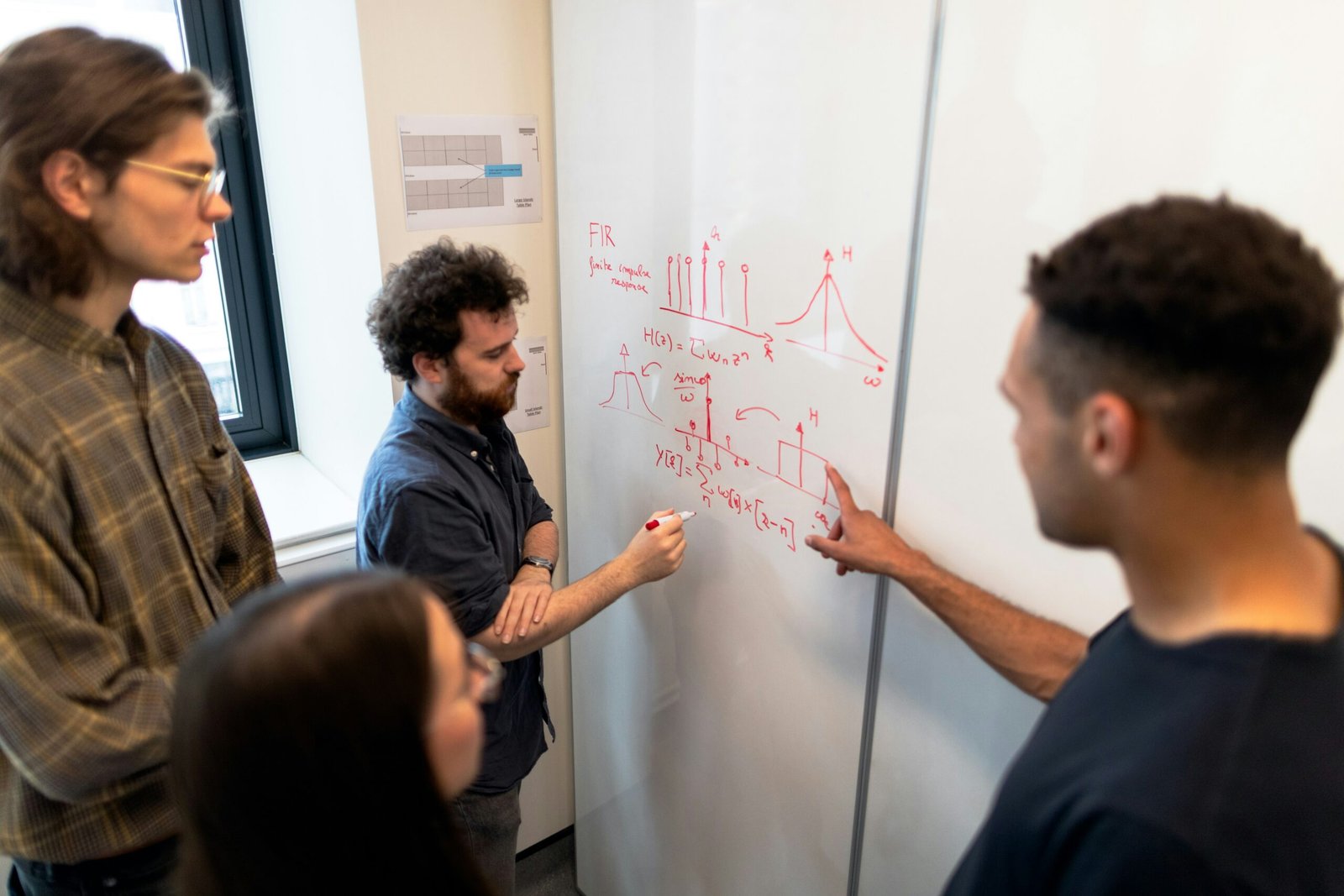 Four colleagues gathered around a whiteboard covered in technical diagrams, working through a problem together